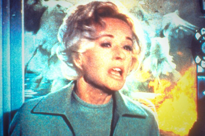 A woman looks alarmed, surrounded by vibrant chaos with blurred bird silhouettes and bright flames in the background.