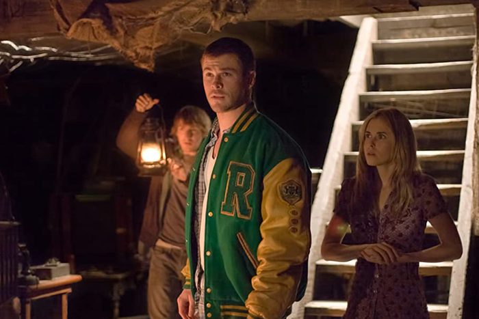 Man wearing a green varsity jacket stands alert; another man holds a lantern, illuminating a woman beside stairs in a dimly lit room.