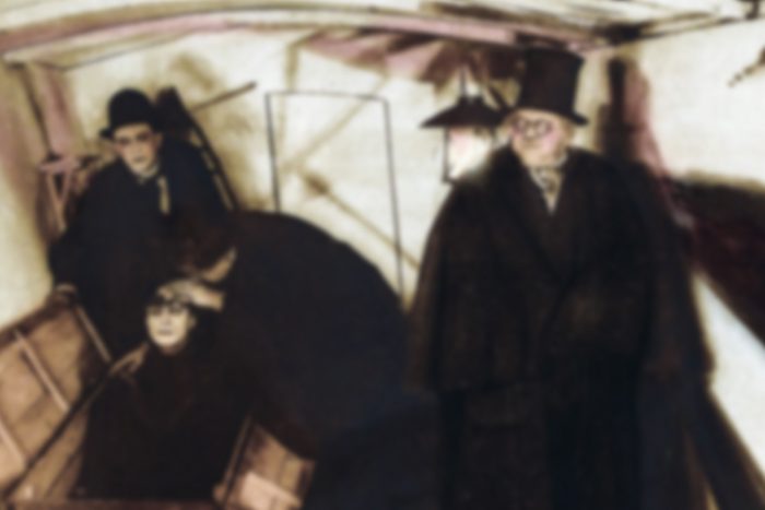 Figures in dark coats stand and sit solemnly inside a dimly lit wooden room, with hats and scarves adding to the atmospheric gloom.