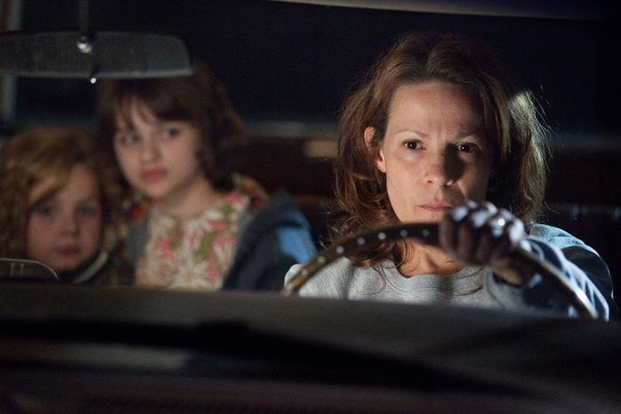 A woman drives a car intently, while two children sit in the back seat, appearing concerned, in a dimly lit setting.