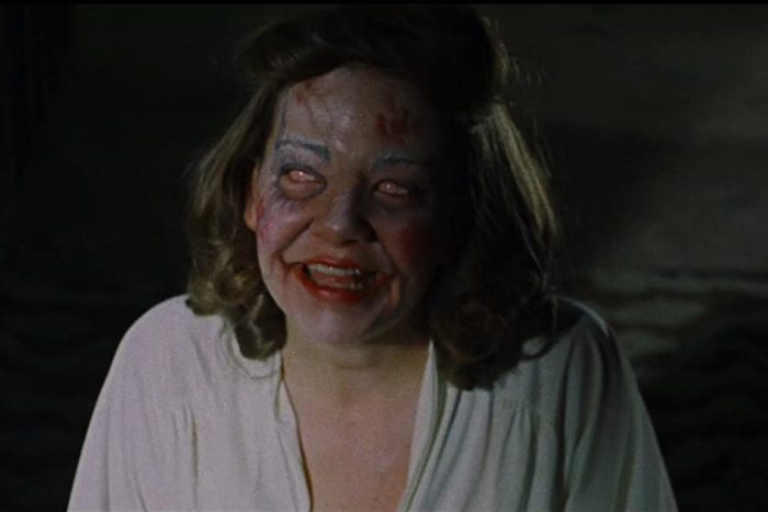 A person, with unsettling makeup, smiles widely in a dimly lit room.