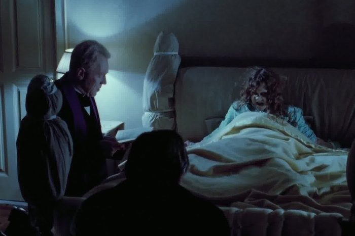 A worried child sits up in bed. Two figures approach with a book in a dimly lit room, conveying a tense atmosphere.