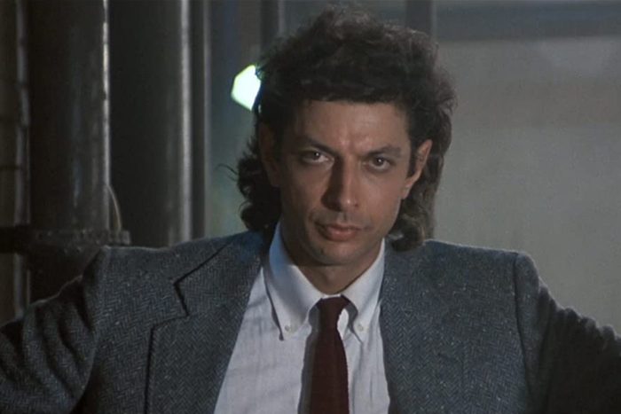 A person in a suit jacket stands confidently, hands on hips, in an industrial setting with pipes and muted lighting.