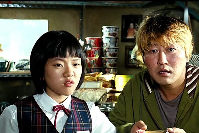 Two people look intently, surrounded by noodle cups and kitchenware in a cozy indoor setting.