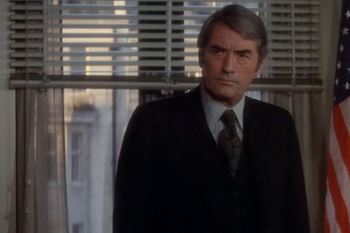 Man stands serious, wearing suit, in office with American flag and window blinds partially open to outside buildings.
