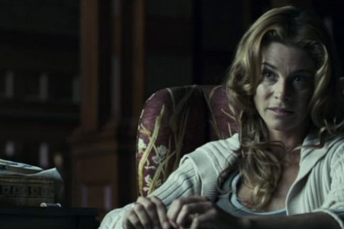 A woman sits in a patterned armchair, looking contemplative, in a dimly lit room with wooden paneling and a table nearby.