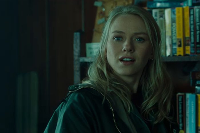 Woman looks towards the viewer, surrounded by books on a shelf, in a dimly lit room.