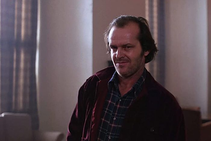 A man stands smiling slightly, wearing a plaid shirt and red jacket, inside a softly lit room with curtains and furniture.