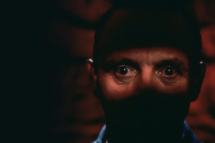 A face partially lit, eyes staring directly, surrounded by shadows and a dark, indistinct background.