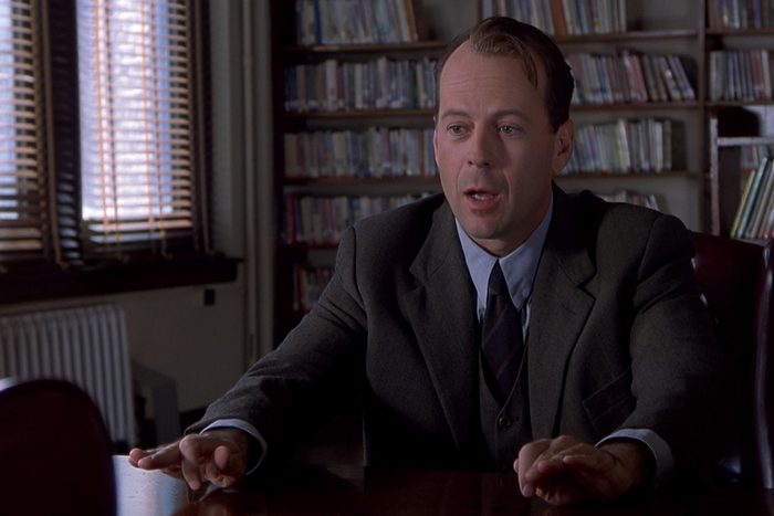 Man in suit speaking at table; background shows bookshelves and window with blinds in a dimly lit room.