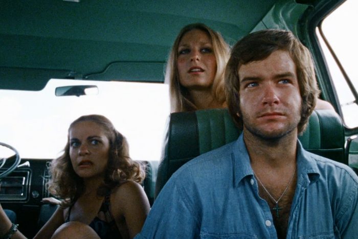 Three people sit in a vehicle, looking out the window with expressions of concern. The interior is dark green.