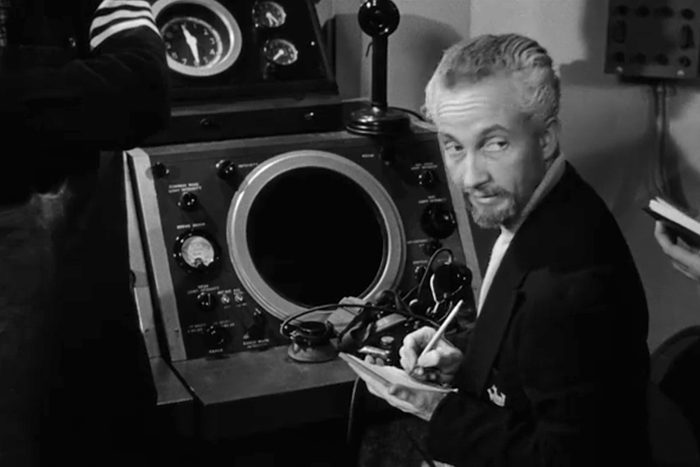 Man writing notes beside vintage radar equipment, looking back, surrounded by dials and switches, in a dimly lit room.