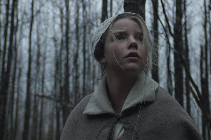 A woman in an old-fashioned cloak looks upwards, standing in a bare, wintry forest.
