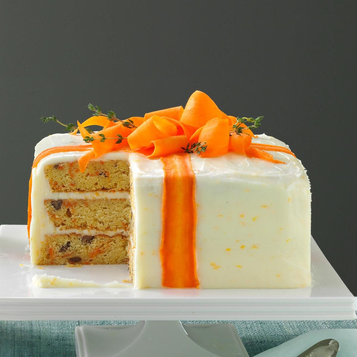 A square carrot cake with white frosting, topped with carrot ribbons and herbs, sits on a white stand in a neutral background.