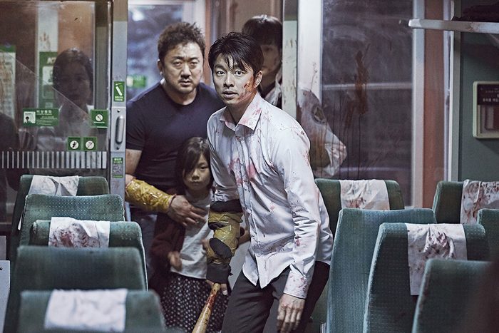 Man with bloody shirt holds weapon, cautiously moving through a train aisle with others, including a child, in tense atmosphere.