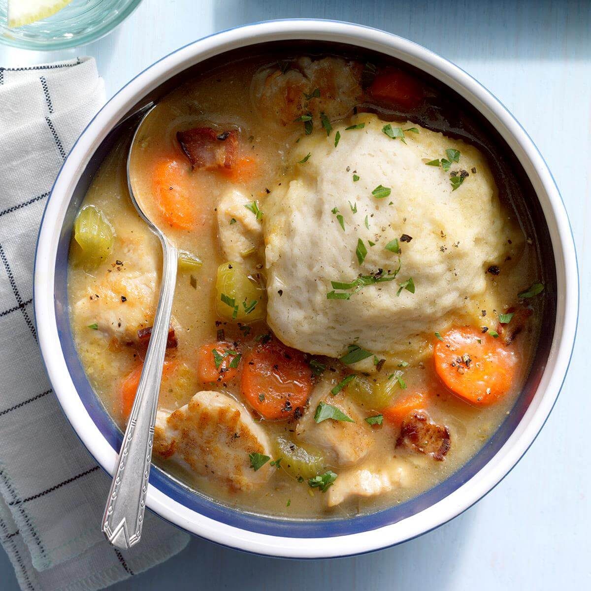 A bowl holds chicken and dumplings topped with herbs, surrounded by sliced carrots and celery, with a spoon placed inside, on a wooden table.