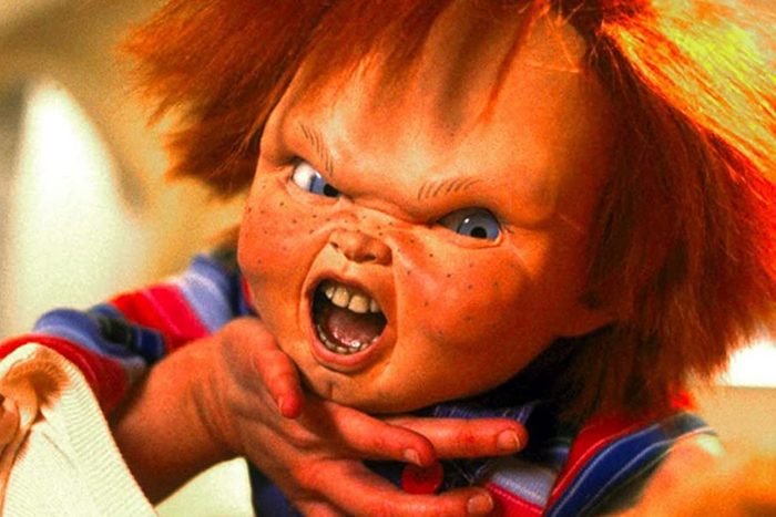 A doll with wild red hair screams angrily, hands gripping something tightly, in a brightly lit room.