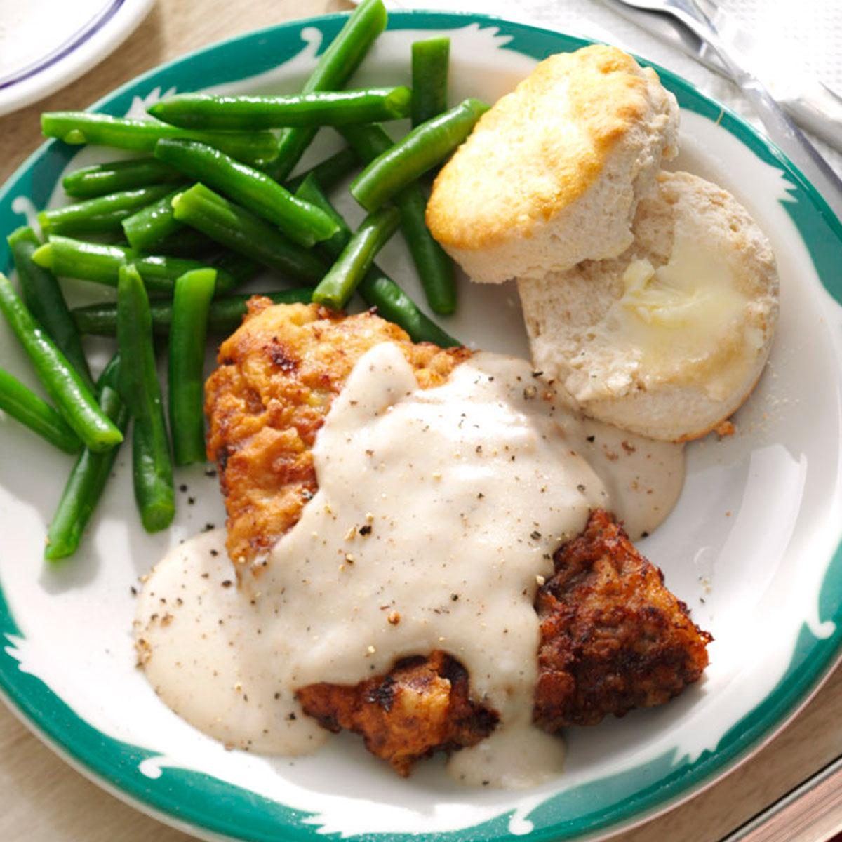 Fried chicken is topped with gravy, accompanied by green beans and a buttered biscuit, on a plate with a green patterned rim.