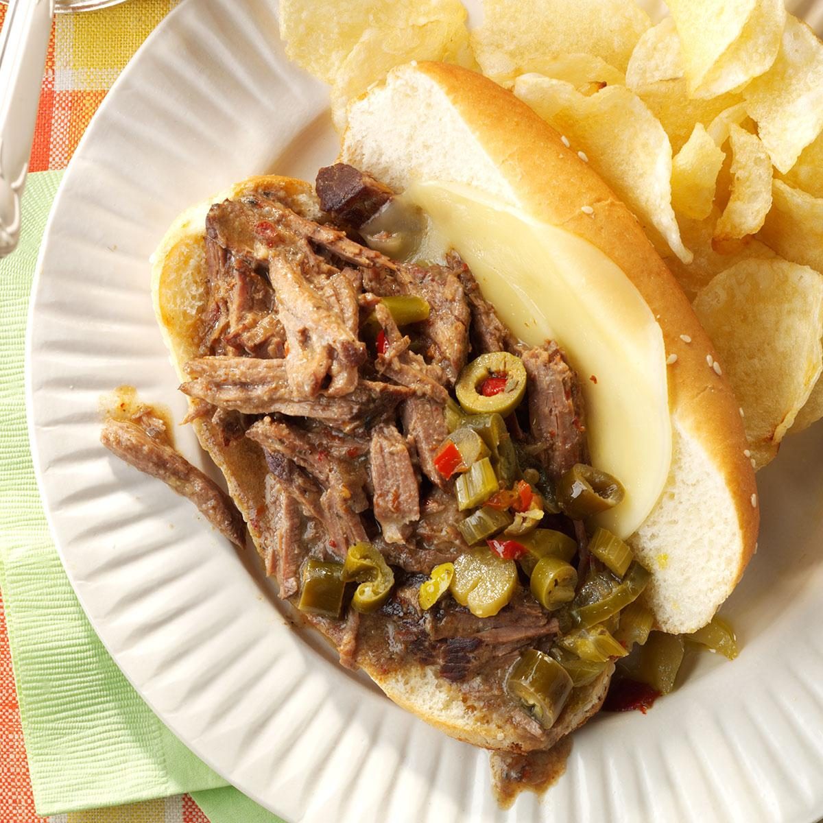 Sandwich with shredded beef and peppers rests on a plate beside potato chips.
