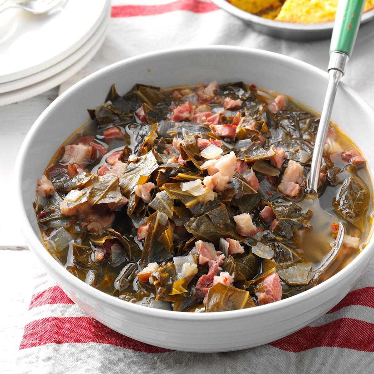 Bowl holds collard greens and bacon bits, sitting in broth with spoon. Set on a table with striped cloth and plates nearby.