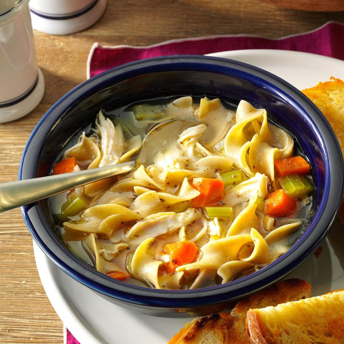 Bowl contains chicken noodle soup with carrots and celery; spoon rests inside. Served on a wooden table with toasted bread alongside.