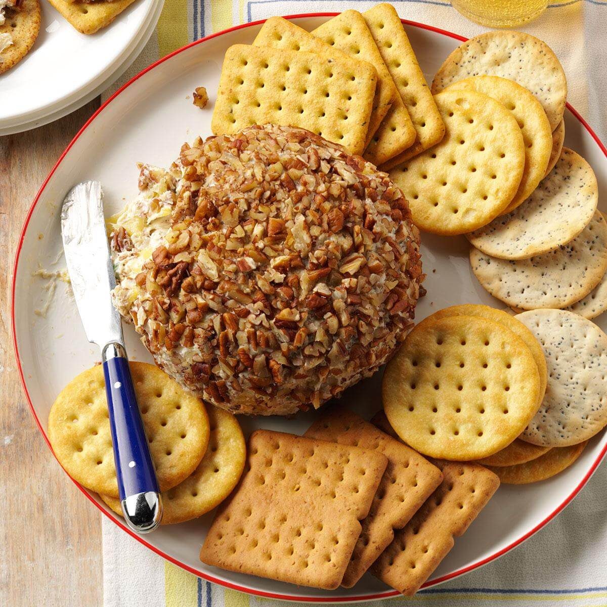 Cheese ball coated in chopped nuts rests on a plate, surrounded by various crackers. A knife with a blue handle is positioned beside it.