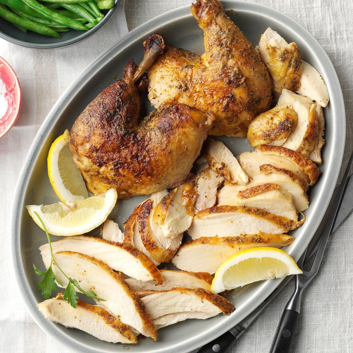 Roasted chicken pieces rest on a gray platter, garnished with lemon wedges and parsley, surrounded by a white cloth and a bowl of green beans.