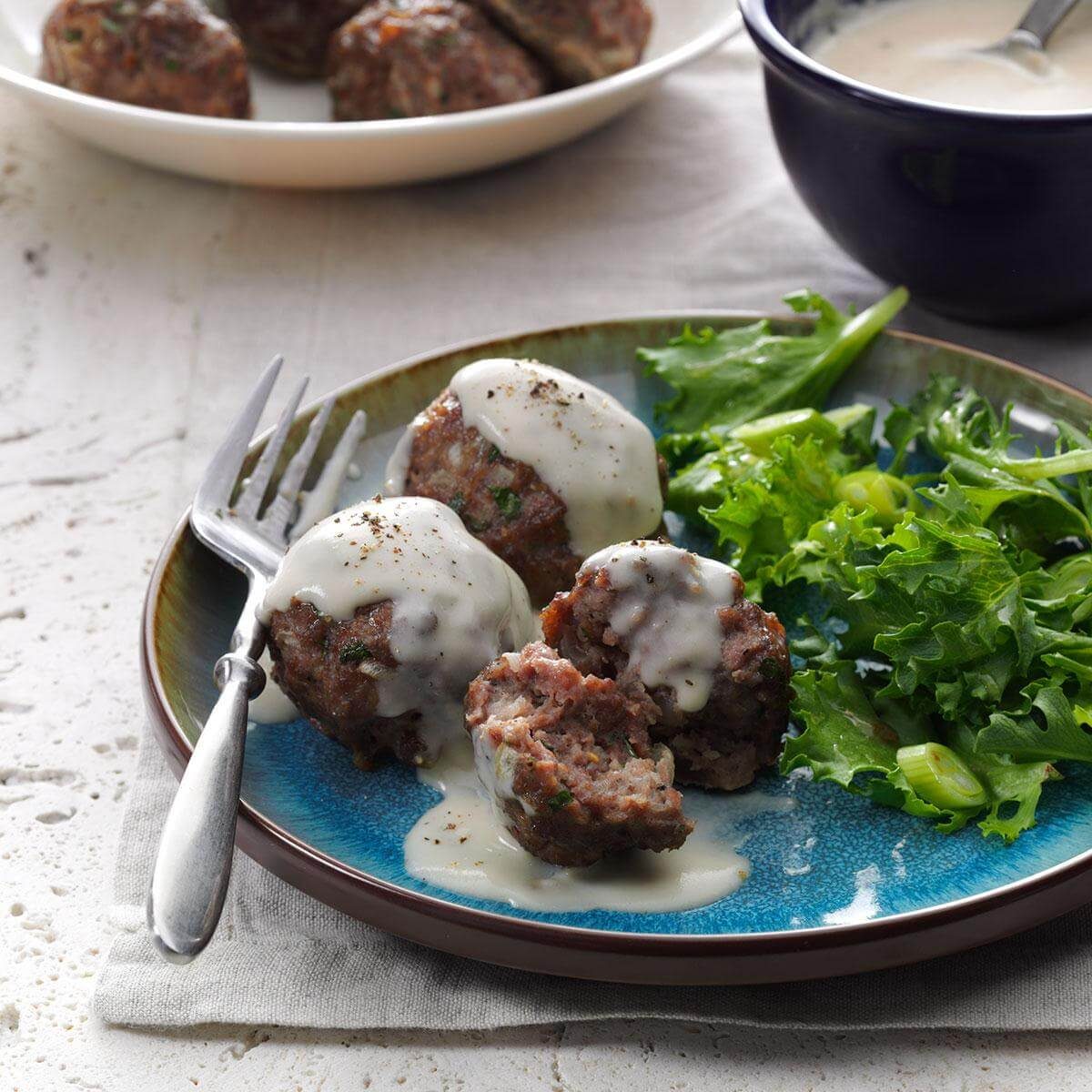 Meatballs covered in white sauce sit beside mixed greens on a blue plate, with a fork resting on the side; bowl of sauce nearby.