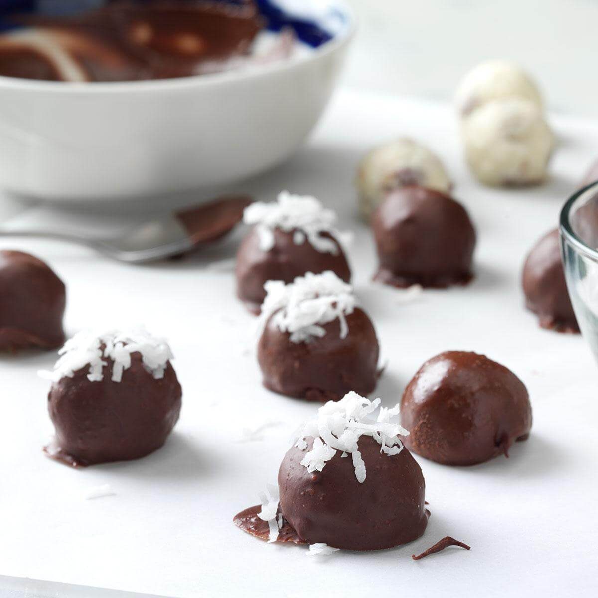 Chocolate coconut truffles rest on white parchment paper, with a bowl and spoon in the background.
