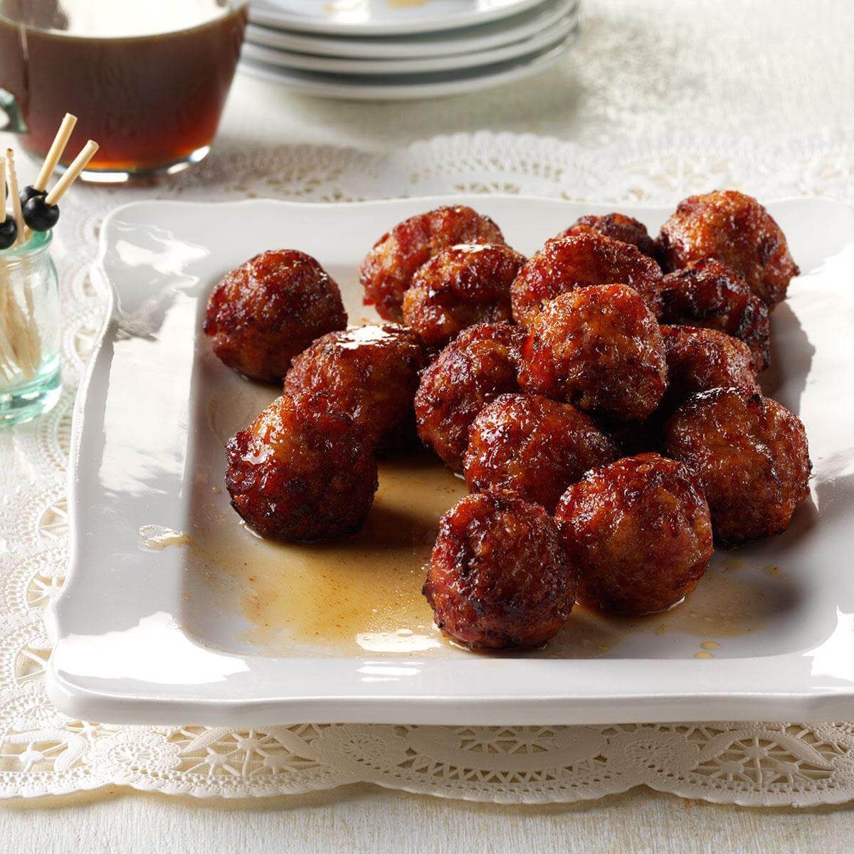 Meatballs glisten on a white square plate, resting on a lace tablecloth with toothpicks and a glass container nearby.