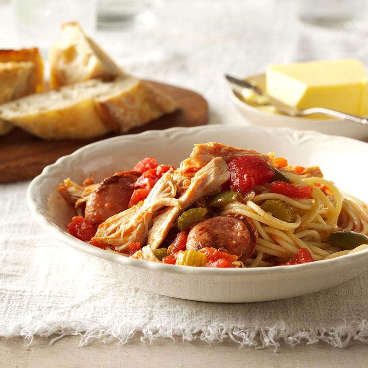 Pasta dish sits in a white bowl, topped with sausage and colorful vegetables. Rustic bread and butter are nearby on a textured tablecloth.