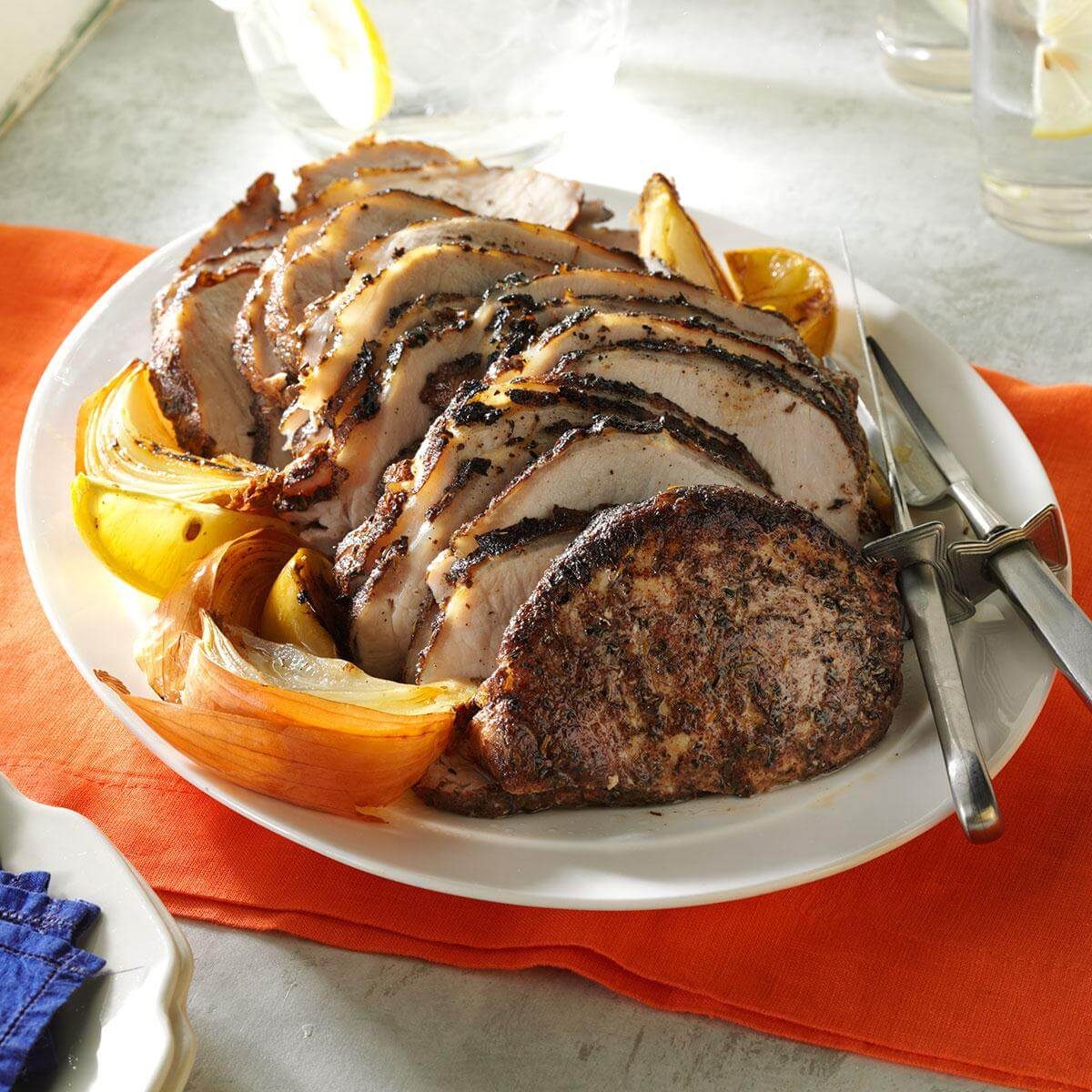 Sliced roast pork sits on a plate, garnished with roasted onions, amidst a bright orange tablecloth setting.