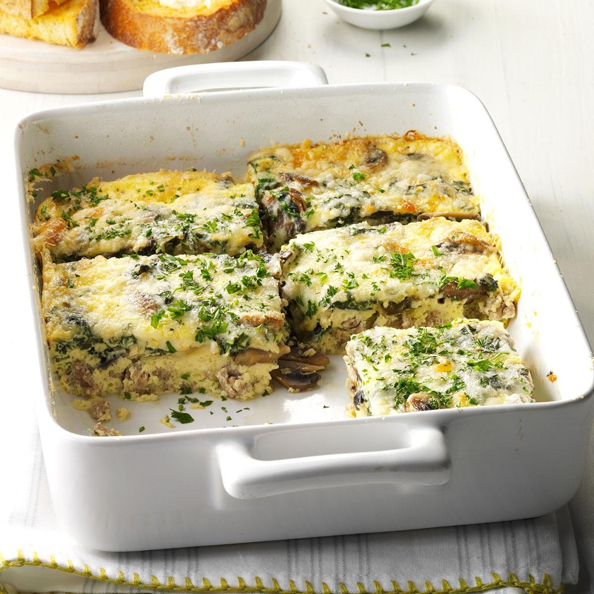 Casserole sits cut into squares in a white baking dish, topped with melted cheese and chopped herbs. Sliced bread is in the background.