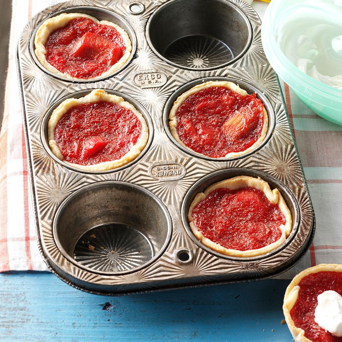 Tartlets fill a vintage muffin tin; red filling contrasts with the metal. Nearby, a dish with white contents and checkered cloth complete the scene.