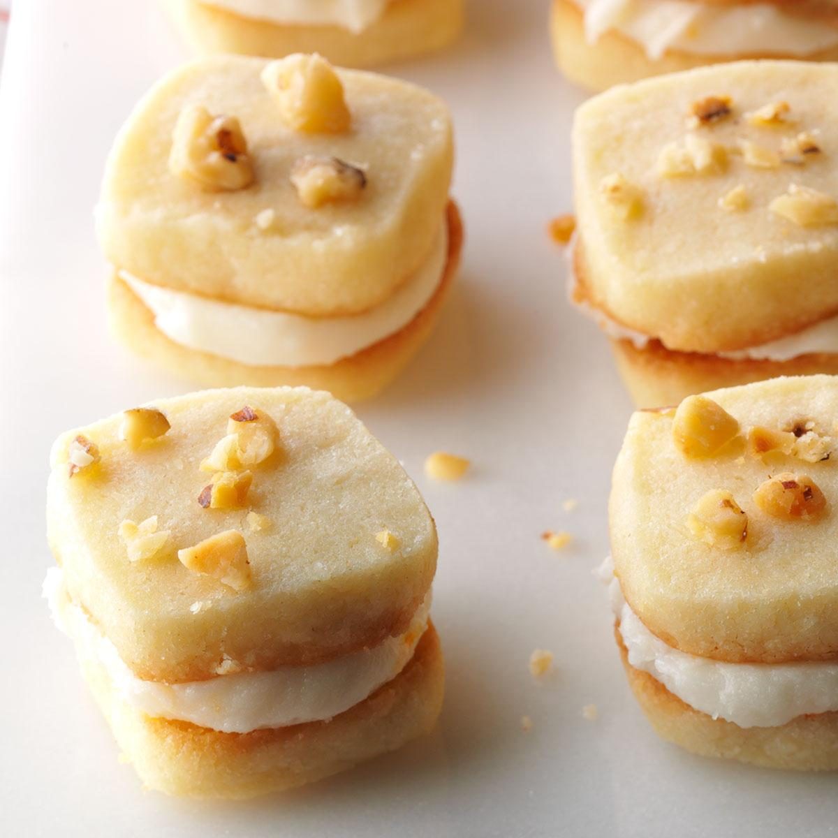 Square cookies layered with cream filling, topped with chopped nuts, rest on a white surface in a minimal arrangement.