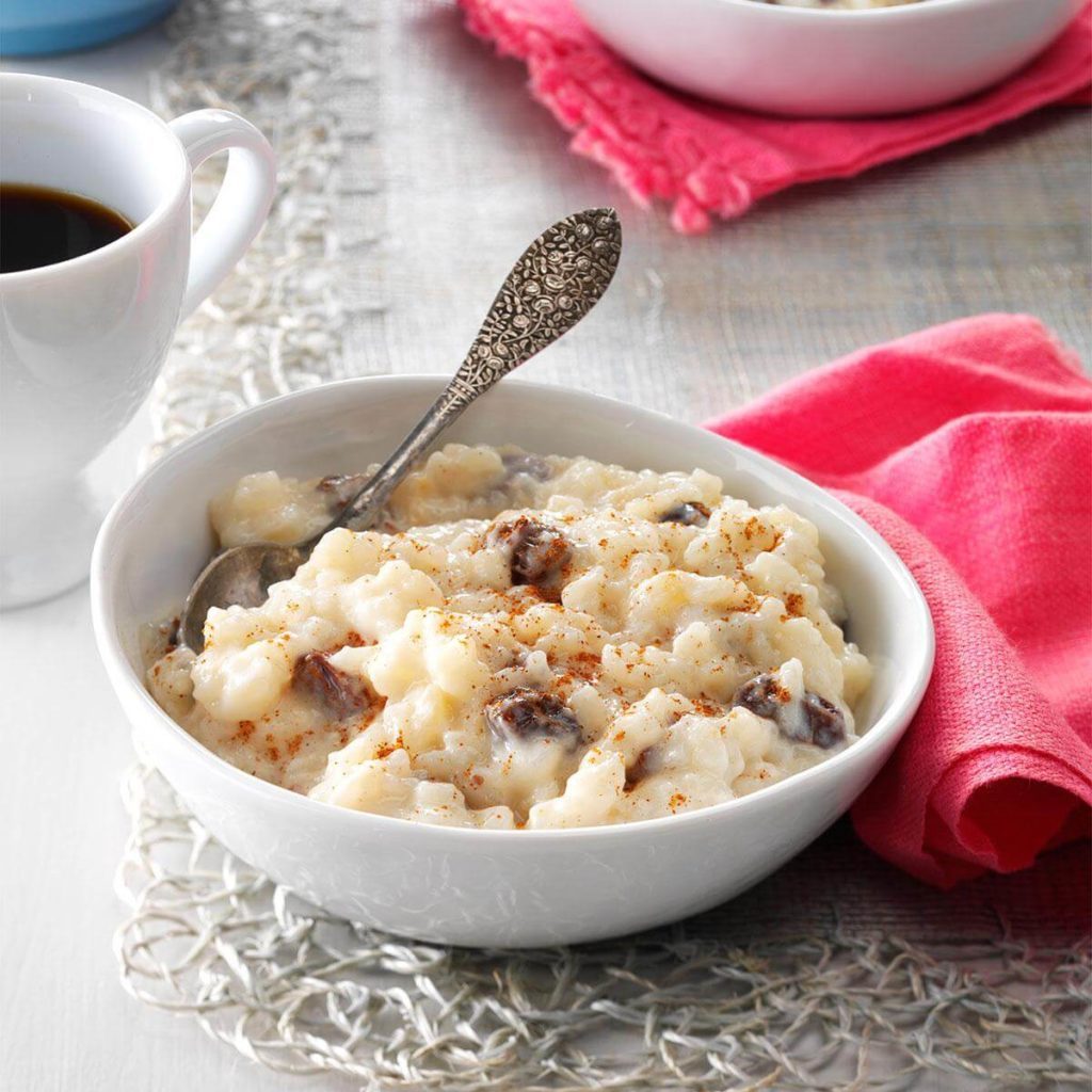 A bowl of rice pudding sits with a spoon, beside a cup of coffee on a textured table with pink napkins.