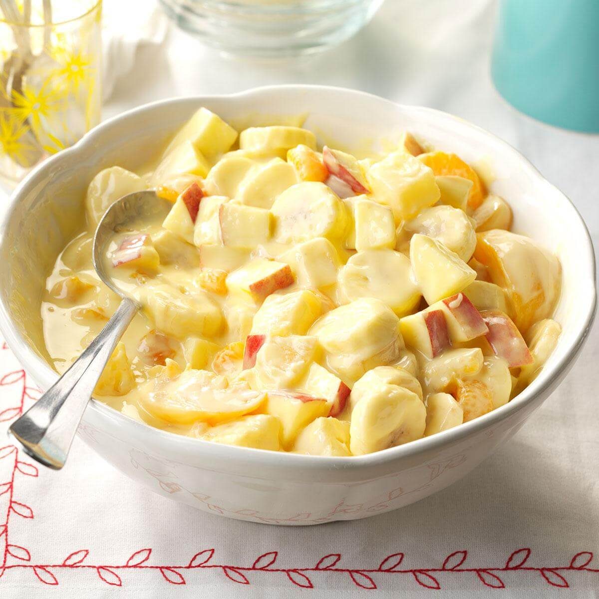 Fruit salad sits in a bowl, mixed with creamy sauce, surrounded by a white tablecloth with red embroidered patterns.