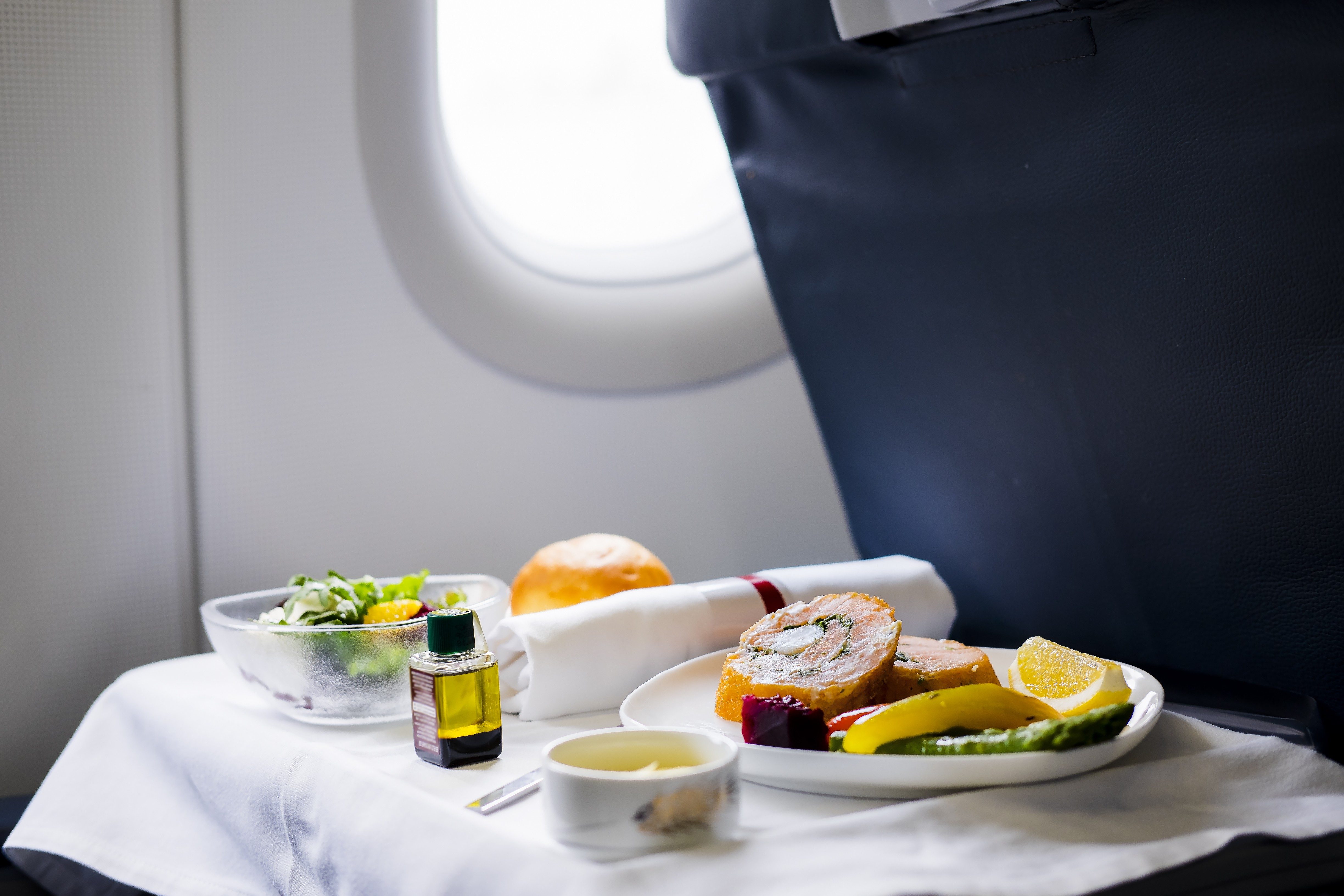 Meal placed on an airplane tray table, featuring a salad, bread roll, oil bottle, and plate of sliced meat and vegetables.
