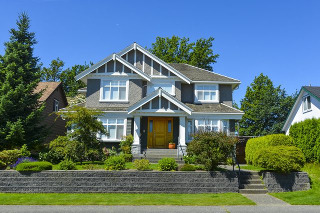 House stands amid manicured bushes and trees under a clear blue sky, with a stone pathway leading to the front door.