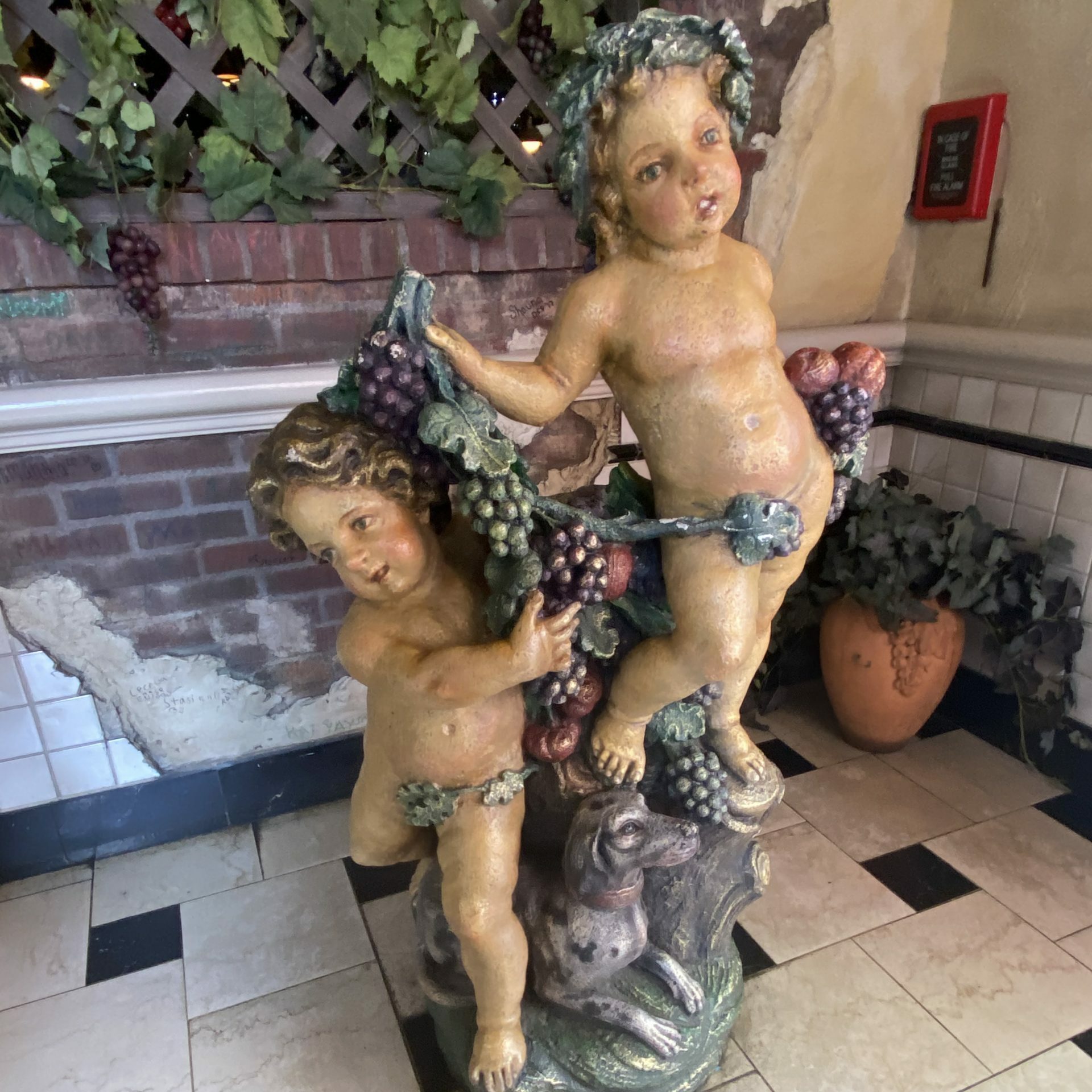 Statue depicts cherubs holding grapevines, with a dog at their feet. Situated indoors on checkered floor, surrounded by ivy and a brick wall.
