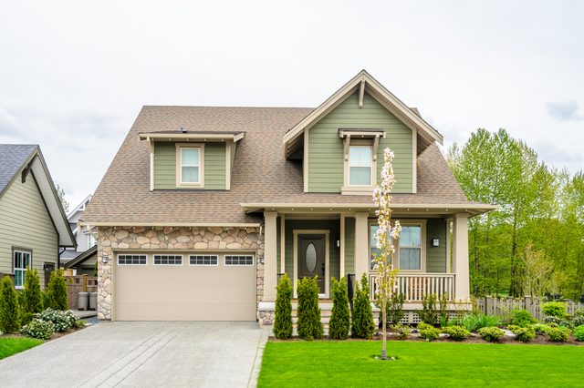 House stands quietly, featuring green siding and stone accents, nestled among trees with a green lawn and driveway in a suburban neighborhood.