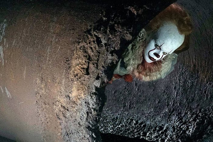 A clown with white face paint peers from a sewer opening, surrounded by dark, textured walls.
