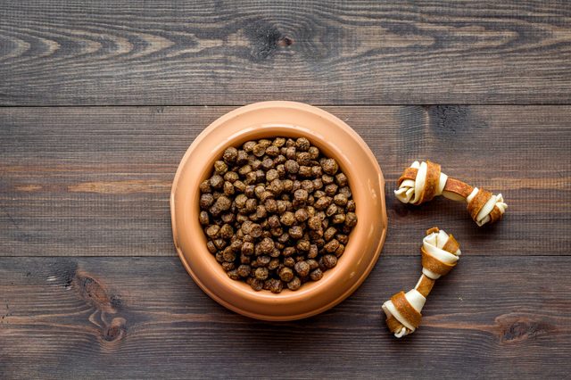 Dog food fills a brown bowl, sitting on a wooden surface with two twisted rawhide bones nearby.