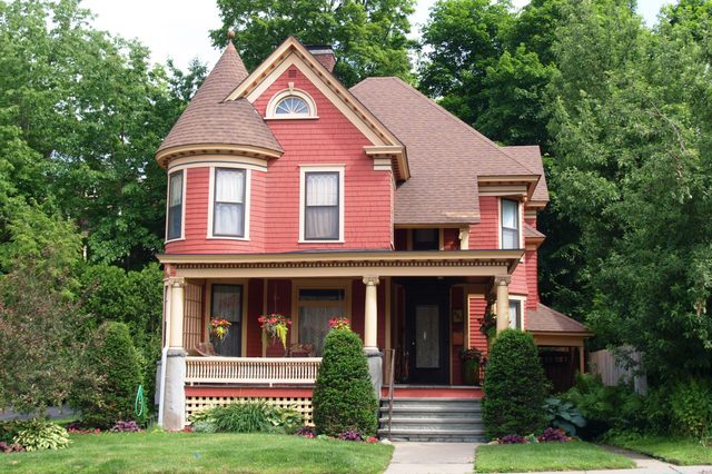 A Victorian-style house stands amid lush greenery, featuring red siding, ornate trim, and a wraparound porch adorned with hanging plants.