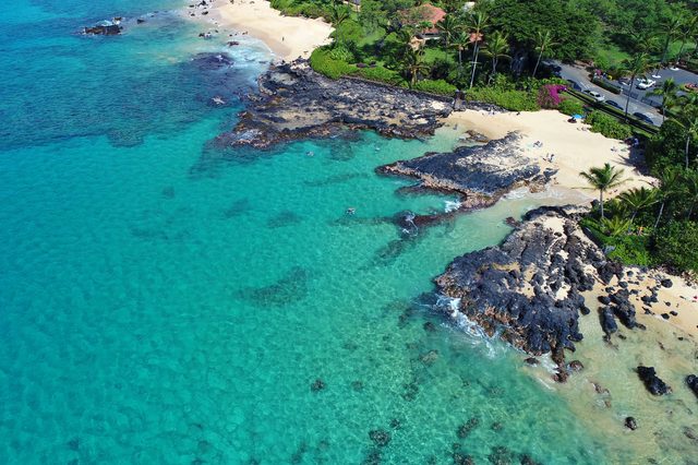 Turquoise ocean waves flow against rocky shoreline and sandy beach, bordered by lush greenery and palm trees, with people sunbathing and swimming nearby.