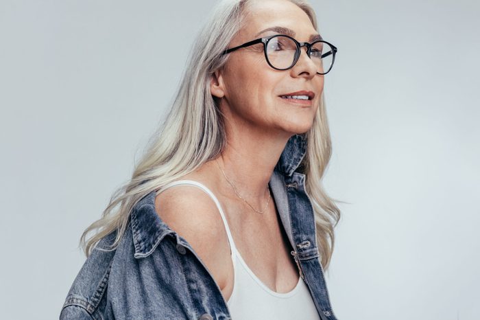 Beautiful senior woman in casuals looking at copy space against grey background. Stylish mid adult caucasian woman in denim shirt and eyeglasses looking away.