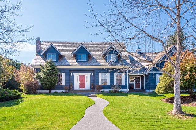 A blue house stands on a lush lawn with a winding path, surrounded by trees and shrubs under a clear, blue sky.