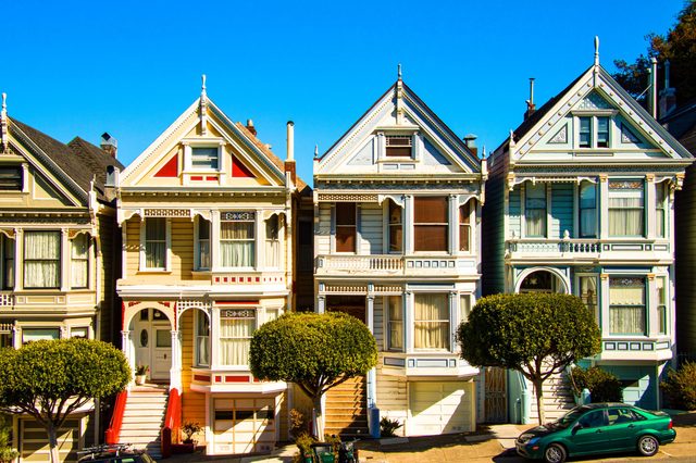 Victorian-style houses stand brightly, showcasing ornate architecture, along a sunny street with neatly trimmed trees and a clear blue sky.