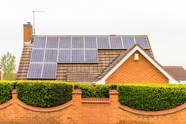 Solar panels cover a sloped roof, bordered by a brick wall and green hedge, under a clear sky.