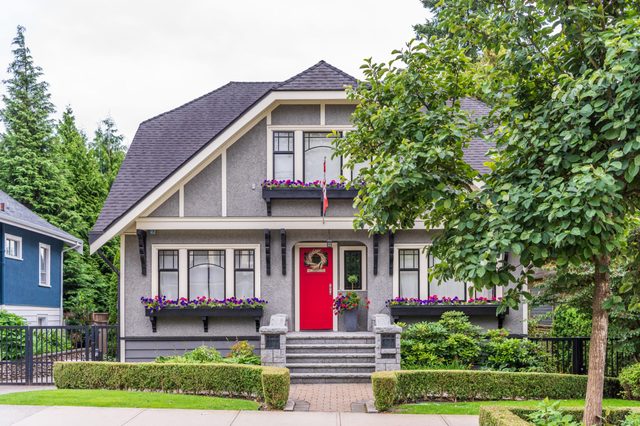 A charming house with a red door stands amidst lush greenery, featuring floral window boxes and a neatly trimmed hedge in front.